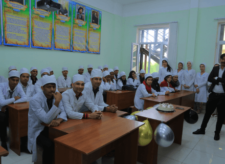 A group of students in white chef uniforms and hats seated at desks in a classroom with a few standing at the back, celebrating with balloons and a cake on a table, organized by Tajhind Edute